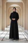 Woman in a black abaya standing in a sunlit hallway with arches.