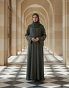Woman in a long dark green Abaya standing in a sunlit hallway with arched columns.