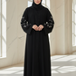 Woman wearing a black abaya with floral embroidery in a bright, sunlit room.