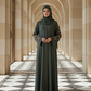 Woman in a long dark green Abaya standing in a sunlit hallway with arched columns.
