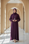 Woman in a long purple dress standing in a sunlit hallway with arches.